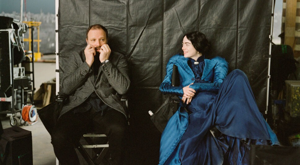 Das Bild zeigt Yorgos Lanthimos und Emma Stone, die auf einem Filmset in Regiestühlen sitzen. Lanthimos zieht eine Grimasse und hält seine Hände vor das Gesicht, während Stone ihn lachend ansieht. Sie trägt ein aufwendiges, blaues Kostüm aus einer historischen Epoche. Der Hintergrund besteht aus schwarzem Stoff und technischem Equipment. Die Szene wirkt entspannt und humorvoll und vermittelt die kreative Atmosphäre, die typisch für Yorgos Lanthimos beste Filme ist.