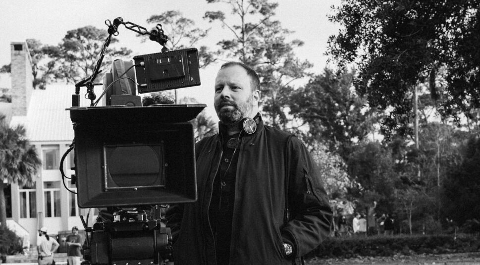 Das Bild aus dem Artikel zu Yorgos Lanthimos' besten Filmen zeigt den griechischen Regisseur hinter einer großen Filmkamera. Er trägt Kopfhörer um den Hals und eine Jacke. Im Hintergrund sind Bäume und ein Haus zu sehen, während einige Menschen im Freien beschäftigt sind. Die Szene wirkt professionell und auf einem Filmset aufgenommen.