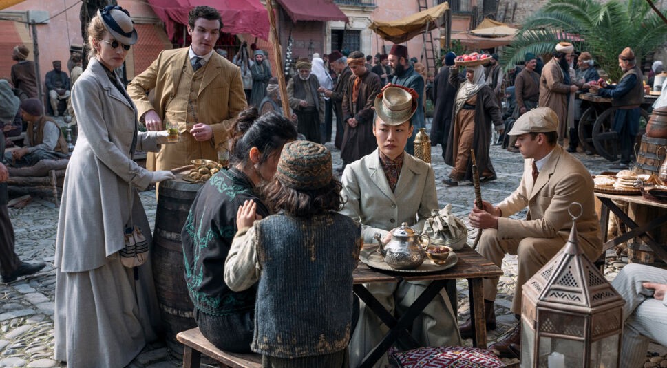 In dieser Szene aus Young Sherlock sind Hero Fiennes Tiffin, Dónal Finn, Zine Tseng und Natascha McElhone in einem historischen Marktplatzsetting zu sehen. Die Figuren tragen detailreiche Kleidung im Stil des späten 19. Jahrhunderts. Zine Tseng sitzt im Zentrum der Szene, umgeben von traditionellen Gegenständen wie einer Teekanne und orientalischen Laternen. Die Atmosphäre ist lebendig und geschäftig.