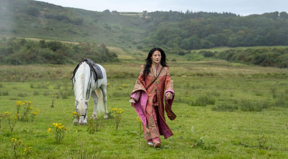 In dieser Szene aus Young Sherlock ist Zine Tseng in einem aufwendig verzierten, rosafarbenen Gewand zu sehen. Sie schreitet über eine weite grüne Wiese, während ein weißes Pferd mit Sattel im Hintergrund grast. Die Landschaft ist ruhig und ländlich, was der Szene eine friedliche, fast mystische Stimmung verleiht.