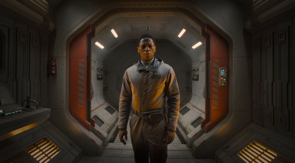 Auf dem Bild in der Kritik zu Alien Romulus siehst Du David Jonsson als Andy in einem grauen Raumanzug in einem futuristischen Korridor stehen, der von hellen Lichtern beleuchtet wird.