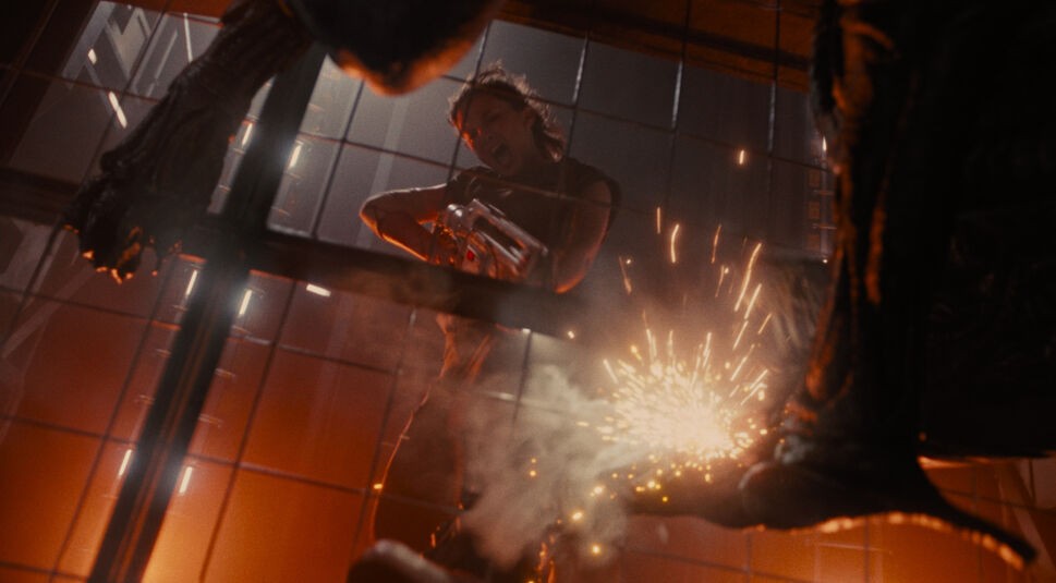 Auf dem Bild in der Kritik zu Alien Romulus siehst du Cailee Spaeny als Rain Carradine in einer intensiven Kampfszene. Sie schießt mit einer Waffe auf das Alien, das außerhalb des Bildes zu sein scheint. Funken fliegen, während die Waffe abgefeuert wird, und der Hintergrund ist in ein bedrohliches rotes Licht getaucht.