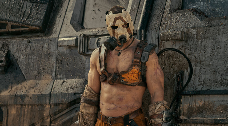 Im Bild zur Borderlands-Filmkritik ist Krieg (gespielt von Florian Munteanu) zu sehen. Er steht vor einem grauen riesigen Objekt, das den gesamten Hintergrund füllt. Es könnte sich um ein großes Auto oder ein massives Tor handeln, dass mit Schlamm bespritzt ist. Kreig selbst trägt eine weiße Maske über seinem Gesicht, die eine Art Sauerstoffventil zu haben scheint. Sein nackter Oberkörper ist zu sehen. Er trägt einen Umschnallgurt über Schultern und Brust. Seine Unterarme sind mit Tüchern umwickelt, an denen Waffengurte oder ähnliches befestigt sind. Seine Arme, seine Brust und sein Bauch sind vernarbt.