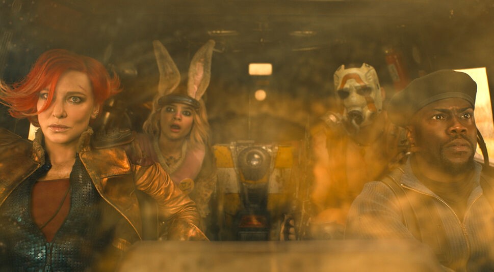 Im Bild zur Borderlands-Filmkritik sind 5 Hauptfiguren zu sehen. Sie sitzen in einem Auto und schauen erschrocken durch die Frontscheibe an der Kamera vorbei. Links im BIld sitzt Lilith (Cate Blanchett) auf dem Beifahrersitz. Sie hat eine rote Kurzhaarfrisur. Auf ihrer Stirn ist eine kleine Schnittnarbe. Sie trägt eine goldbraune Lederjacke und ein rotes Oberteil darunter. Auf dem Sitz hinter ihr sitzt Tiny Tina (Ariana Greenblatt). Ihr Mund ist offen. Sie trägt einen schwarzen Haarreif an ihrer Stirn, an dem große plüschige Hasenohren befestigt sind. Neben ihr sitzt der Roboter Claptrap auf der Rückbank. Er ist also zentral im Bild. Er ist Kastenförmig und hat ein Kameraauge in der Mitte. Der Kasten ist gelb-weiß gestreift. Rechts von ihm im Bild sitzt ebenfalls auf der Rückbank Krieg (Florian Munteanu). Er trägt eine Art Sauerstoffmaske in Weiß über sein gesamtes Gesicht. Vor ihm auf dem Fahrersitz ist Roland (Kevin Hart) zu sehen. Er trägt eine Soldatenkappe in Schwarz und eine dunkelblaue Jacke. Es sieht aus als wäre die Aufnahme durch die Forntscheibe des Fahrzeugs gemacht worden, die staubig ist.