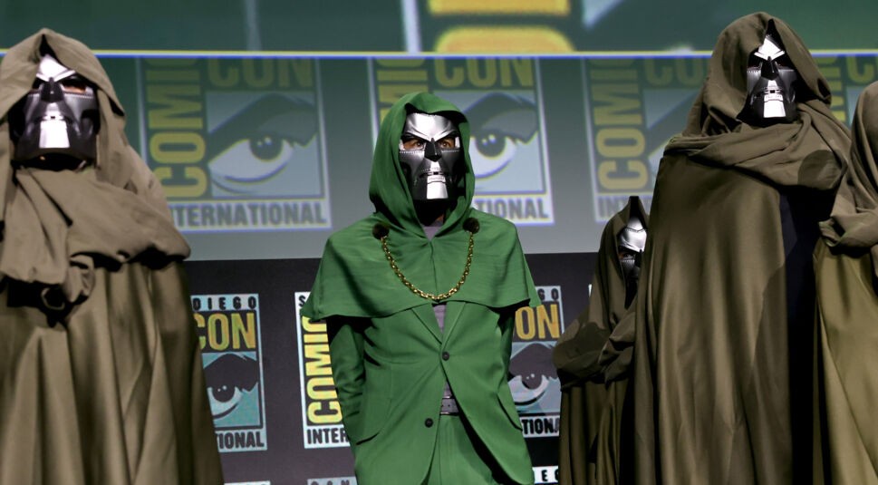 Auf dem Bild von der San Diego Comic Con 2024 steht Robert Downey Jr. in einem Doctor-Doom-Kostüm in der Mitte. Er trägt eine grüne Kapuze, einen Umhang und eine silberne Maske. Um ihn herum stehen weitere Personen in dunkelgrünen Doctor-Doom-Kostümen. Im Hintergrund ist das Comic-Con-Logo gut sichtbar.