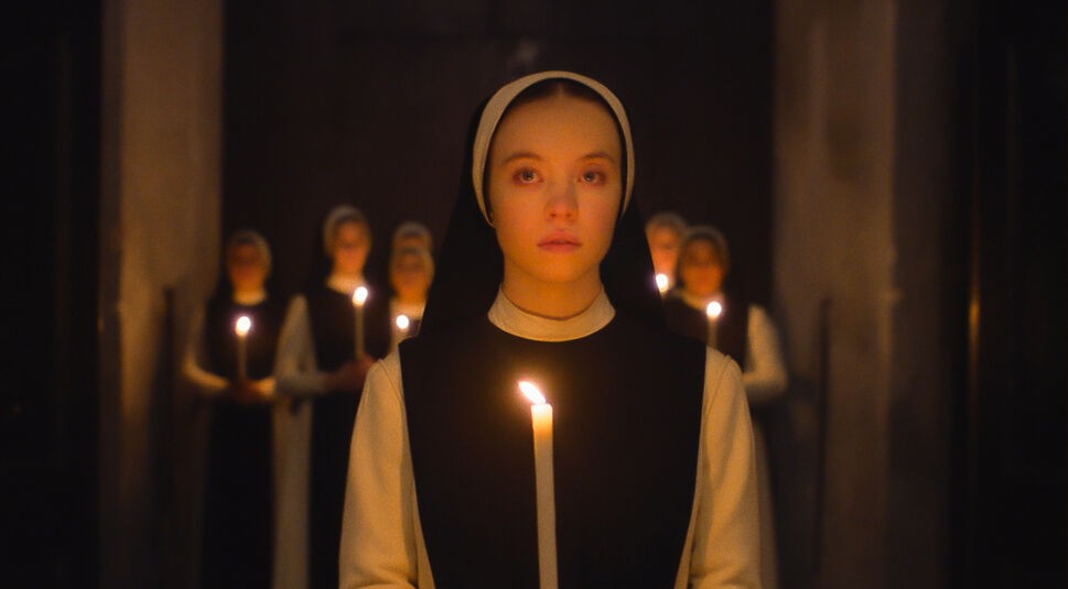 Sydney Sweeney spielt in Immaculate eine Nonne. Sie ist in schwarz-weißer Nonnen-Kutte zentral im Bild zu sehen. Sie hält eine weiße, angezündete Kerze in ihrer Hand und befindet sich in einem dunklen Raum. Im Hintergrund sind verschwommen weitere Nonnen zu sehen, die ebenfalls jeweils eine Kerze in den Händen halten,