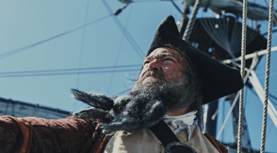 Auf diesem Bild zu den GigaTV Highlights im August ist eine Szene aus der Dokumentation "Piraten: Die Geschichten hinter den Legenden" zu sehen. Ein Pirat mit einem dichten, grauen Bart und einem markanten, wettergegerbten Gesichtsausdruck blickt entschlossen in die Ferne. Er trägt einen klassischen Dreispitz und historische Kleidung, die zu seiner rauen Erscheinung passt. Im Hintergrund sind Teile eines Segelschiffs und Taue zu sehen, was die maritime Atmosphäre der Szene unterstreicht. Die Darstellung wirkt authentisch und fesselnd, passend zur historischen Thematik der Dokumentation.