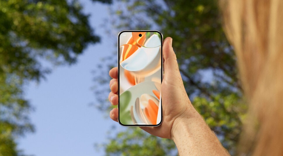 Auf dem Bild hält eine Person das Pixel 9 Pro aus der Serie der neuen Pixel Smartphones in der Hand. Das Smartphone zeigt einen farbenfrohen, abstrakten Bildschirmhintergrund in Orange-, Grün- und Beigetönen. Im Hintergrund sind verschwommene Bäume und ein klarer blauer Himmel zu sehen, was auf eine Nutzung im Freien hinweist.