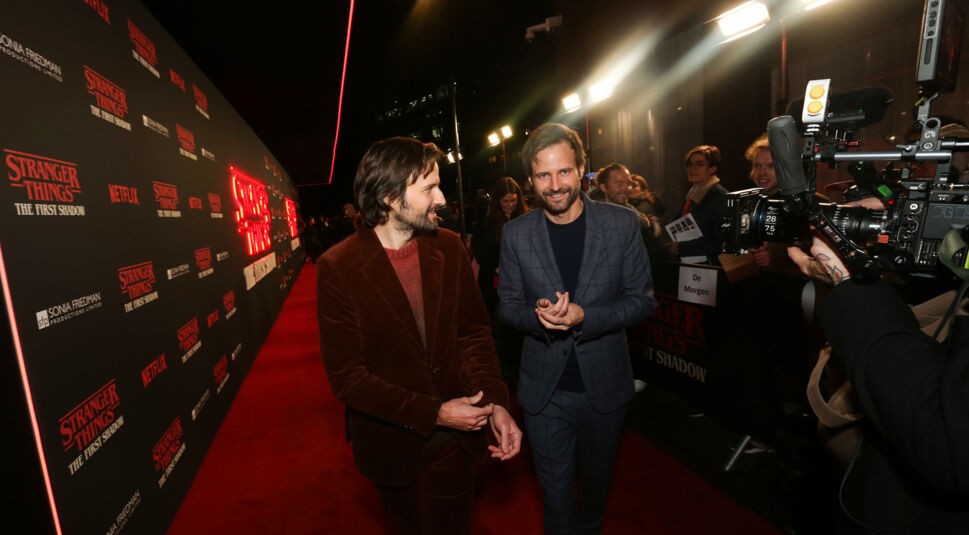 Auf dem Bild für Something Very Bad Is Going To Happen sind Matt und Ross Duffer, die Schöpfer von Stranger Things, auf dem roten Teppich des Theaterstücks "Stranger Things: The First Shadow" zu sehen. Matt Duffer trägt einen braunen Samtanzug, während Ross Duffer in einem grauen, karierten Anzug erscheint. Beide lächeln und unterhalten sich, während sie den Veranstaltungsort betreten. Der Hintergrund ist mit dem Stranger Things Logo und dem Titel der neuen Produktion dekoriert, und das Bild ist von Kameraleuten und Fans umrahmt.