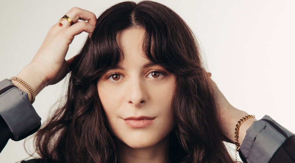 Auf diesem Bild, das als Promo für Something Very Bad Is Going To Happen verwendet wird, ist Haley Z. Boston zu sehen. Sie hat dunkles, welliges Haar und blickt direkt in die Kamera. Ihre Hände sind in einer nachdenklichen Pose an ihrem Kopf platziert, während sie ein sanftes Lächeln auf den Lippen trägt. Die Atmosphäre des Bildes ist ruhig, mit einem schlichten, hellen Hintergrund, der den Fokus auf ihr Gesicht und ihre goldenen Schmuckstücke lenkt.