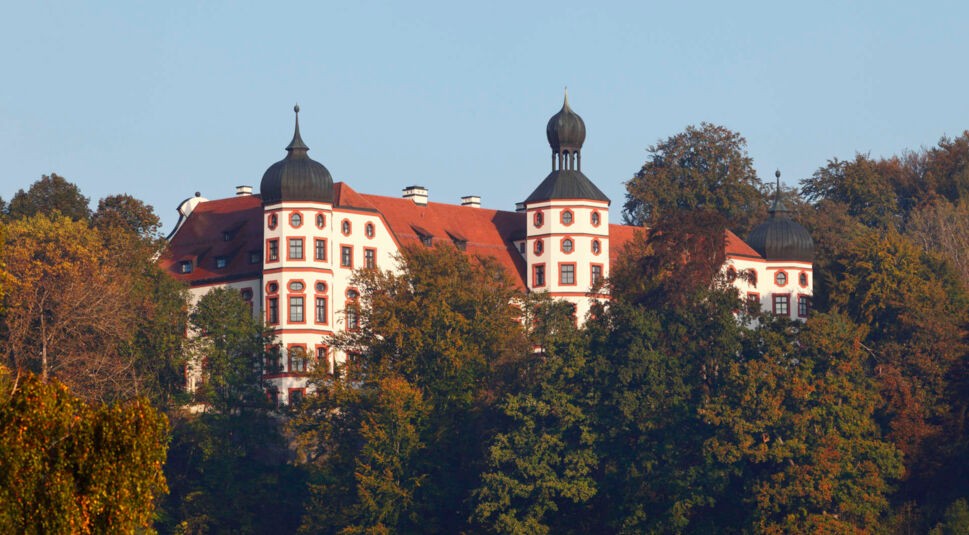 Ein weißes Schloss mit rot gerahmten Fenstern und rotem Ziegeldach ragt aus einem Wald heraus.