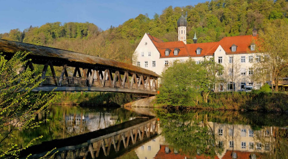 Eine überdachte Holzbrücke führt über einen ruhigen Fluss, im Hintergrund ein beigefarbenes Kirchengebäude mit rotem Ziegeldach