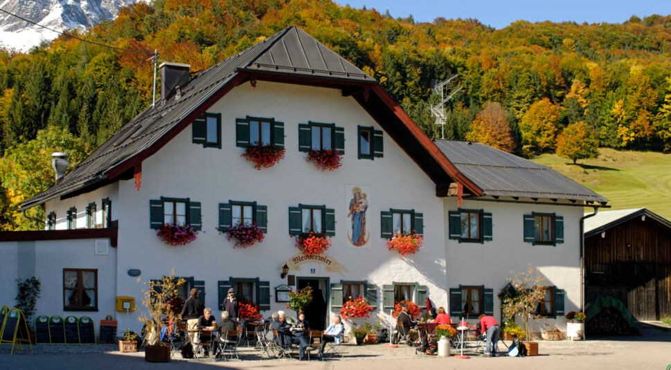 Ein großes weißes Wirtshaus vor einem Wald und Alpenpanorama