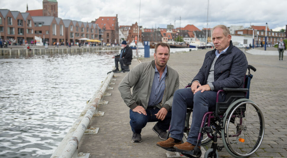 Zwei Männer vor einer Hafenkulisse: einer sitzt im Rollstuhl, der andere kniet neben ihm.