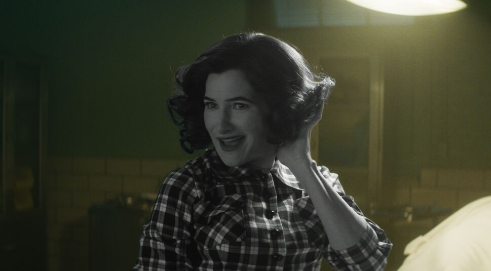 Auf dem Artikelbild zum Agatha Harkness-Artikel ist Agatha Harkness, gespielt von Kathryn Hahn, in einem Retro-Look zu sehen. Sie trägt ein kariertes Hemd und ihre Haare sind in weichen Locken frisiert, die an die 1950er-Jahre erinnern. Ihr Gesichtsausdruck ist schelmisch und sie lächelt verschmitzt, während sie mit einer Hand ihre Haare zurückstreicht. Die Szene ist in Schwarz-Weiß gehalten, was einen nostalgischen und zugleich mysteriösen Charme verleiht. Der Hintergrund ist unscharf, wodurch der Fokus auf Agathas Ausdruck und Pose liegt.