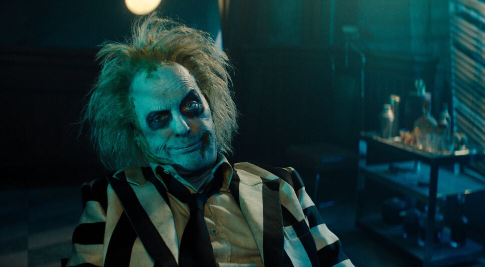 Auf dem Bild zu "Beetlejuice Beetlejuice Ende erklärt" ist Michael Keaton als Beetlejuice in seinem charakteristischen schwarz-weiß gestreiften Anzug zu sehen. Sein Gesicht ist mit dunklem Make-up geschminkt, die Haare sind wild zerzaust. Er sitzt lässig zurückgelehnt, mit einem leicht schelmischen und verschmitzten Lächeln, das seinen exzentrischen und frechen Charakter unterstreicht. Im Hintergrund steht ein Tisch mit verschiedenen Gegenständen, der Raum ist in düsterem, grünlichem Licht gehalten, was eine geheimnisvolle und leicht unheimliche Atmosphäre erzeugt.