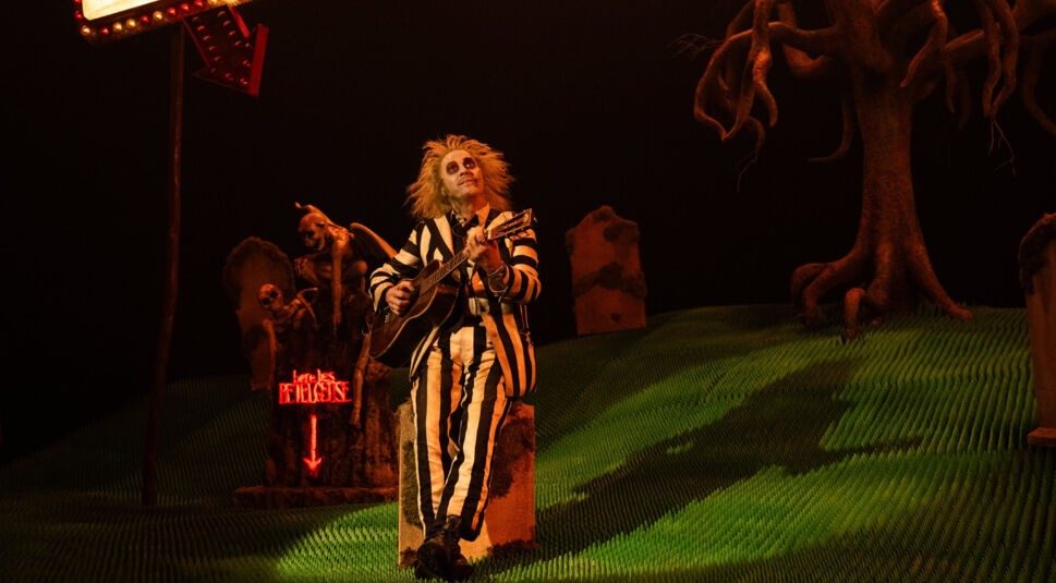 Auf diesem Bild zur Beetlejuice Beetlejuice Kritik spielt Michael Keaton als Beetlejuice Gitarre und sitzt auf einem Grabstein inmitten einer düsteren Friedhofsszene. Er trägt sein charakteristisches schwarz-weiß gestreiftes Outfit, und seine wild abstehenden Haare unterstreichen seine skurrile Erscheinung. Im Hintergrund sind Gräber und eine leuchtende rote Leuchtschrift mit der Aufschrift "Here Lies Beetlejuice" zu sehen. Ein knorriger, kahler Baum steht rechts, und die gesamte Szene wirkt surreal und makaber.