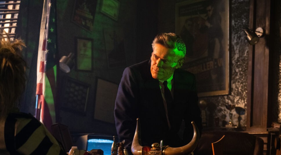 Auf diesem Bild aus Beetlejuice Beetlejuice ist Willem Dafoe als eine mysteriöse Figur mit einer halb freigelegten Gehirnpartie zu sehen. Er lehnt sich auf einen Schreibtisch, während er mit ernstem Blick jemandem außerhalb des Bildes gegenübersteht. Im Raum sind dunkle Wände, eine US-Flagge und ein Poster an der Wand zu erkennen, was der Szene eine düstere, bedrückende Atmosphäre verleiht. Im Vordergrund ist der Kopf von Beetlejuice (Michael Keaton) leicht unscharf zu sehen.