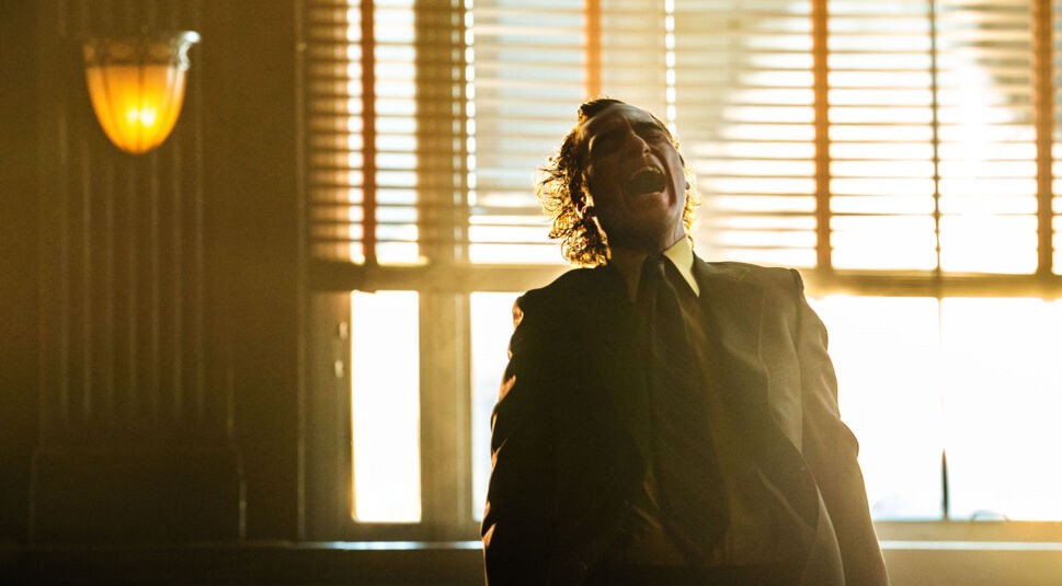 Auf dem Bild zum Joker-Faktenartikel ist Joaquin Phoenix als der Joker in einer dramatischen Szene zu sehen. Er trägt einen Anzug und steht vor einem Fenster mit halb geschlossenen Jalousien. Im goldenen Licht wirkt seine Körperhaltung intensiv, während er den Kopf nach hinten neigt und den Mund weit aufreißt, als würde er schreien oder lachen. Die Stimmung ist düster und spannungsgeladen, typisch für den Charakter des Jokers.