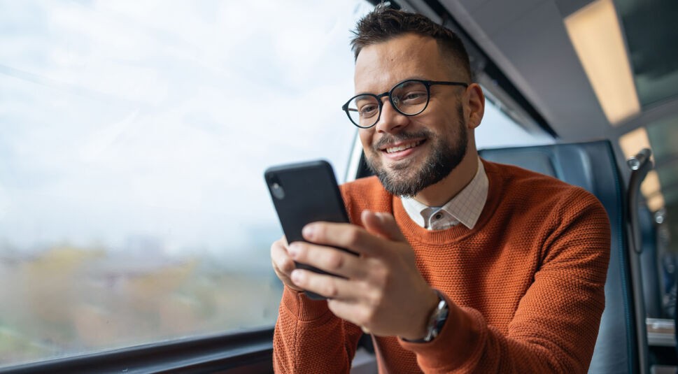 Auf dem Bild in dem Artikel zu den GigaMobil-Tarifen sitzt ein junger Mann mit Brille und Bart in einem Zug am Fenster. Er trägt einen orangefarbenen Pullover und lächelt, während er auf sein Smartphone schaut.