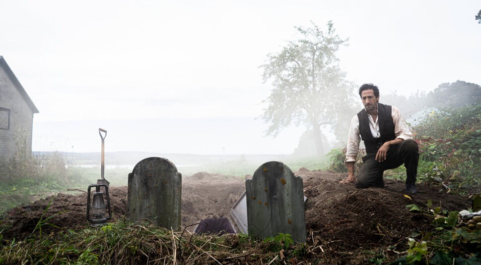 Auf dem Bild zu den GigaTV Highlights im Oktober ist Adrien Brody als Charles Boone in einer düsteren Szene aus "Chapelwaite" zu sehen. Er kniet neben einem frisch ausgehobenen Grab zwischen zwei verwitterten Grabsteinen. Eine Schaufel und eine Laterne liegen im Vordergrund. Der Nebel und die graue Landschaft verstärken die unheilvolle Atmosphäre, während ein einsames Haus im Hintergrund zu sehen ist. Brody trägt zerknitterte, historische Kleidung, und sein Gesichtsausdruck wirkt angespannt und nachdenklich.