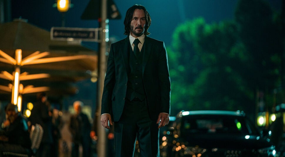 Auf diesem Bild zur "John Wick-Serie Under the High Table" steht Keanu Reeves als John Wick in einem dunklen Anzug auf einer nächtlichen Straße. Die Szene ist in einem städtischen Umfeld angesiedelt, beleuchtet von warmen Straßenlichtern, die einen intensiven und entschlossenen Ausdruck auf seinem Gesicht hervorheben. Im Hintergrund sind unscharfe Personen und geparkte Autos zu erkennen, was die bedrohliche Atmosphäre verstärkt.