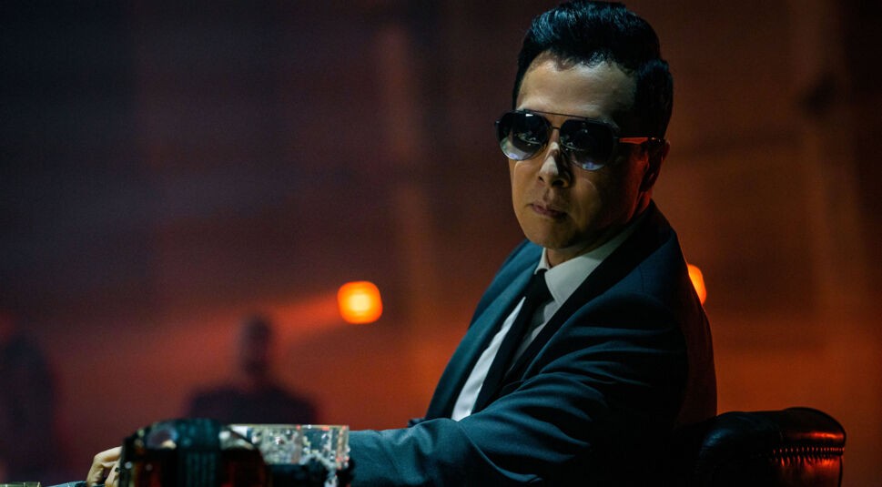 Auf diesem Bild zur "John Wick-Serie Under the High Table" ist Caine, gespielt von Donnie Yen, zu sehen. Er trägt einen eleganten schwarzen Anzug und eine dunkle Sonnenbrille, was seinem Charakter eine coole und gefährliche Ausstrahlung verleiht. Die düstere Beleuchtung im Hintergrund und die orangefarbenen Lichter erzeugen eine bedrohliche Atmosphäre, während Caine konzentriert und entschlossen wirkt. Sein selbstbewusster Gesichtsausdruck deutet auf seine Rolle als ernstzunehmender Gegner hin.