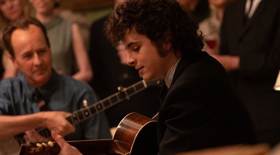 Auf dem Bild zu Like A Complete Unknown ist Bob Dylan, gespielt von Timothée Chalamet, in einer intimen Szene zu sehen, wie er auf einer Akustikgitarre spielt. Umgeben von einer kleinen Gruppe von Menschen, die ihm konzentriert zuhören, wirkt die Atmosphäre nostalgisch und konzentriert. Dylan trägt einen eleganten Anzug, während er in Gedanken versunken zu spielen scheint.