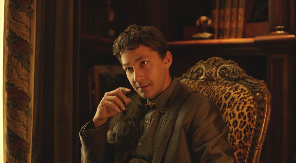 Auf dem Artikelbild von Megalopolis-Kritik ist der Charakter Clodio, gespielt von Shia LaBeouf, in einer intimen Szene zu sehen. Er sitzt in einem kunstvoll verzierten Sessel mit Leopardenmuster, hält eine Zigarette in der Hand und wirkt nachdenklich. Das warme, goldene Licht und die opulente Einrichtung, mit schweren Vorhängen und Bücherregalen im Hintergrund, unterstreichen eine luxuriöse, fast nostalgische Atmosphäre. Clodios Gesichtsausdruck vermittelt eine Mischung aus Selbstbewusstsein und subtiler Unruhe.