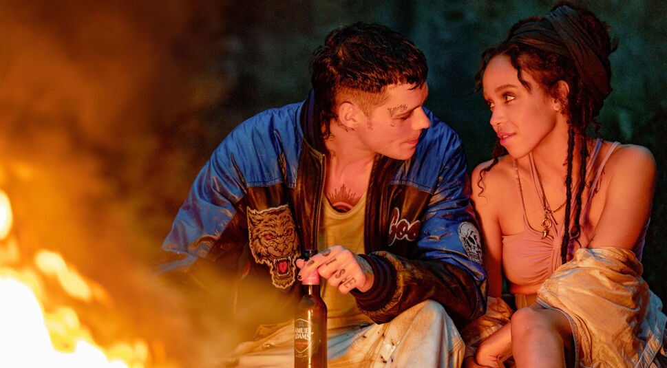 Auf dem Artikelbild zur The Crow-Kritik sind Bill Skarsgård und FKA Twigs in einer intimen Szene am Lagerfeuer zu sehen. Bill trägt eine blaue Jacke mit Tiger-Aufnäher und hält eine Bierflasche in der Hand, während er FKA Twigs liebevoll ansieht. Sie trägt ein rosa Oberteil und eine lockere Jacke, ihre Haare sind in Zöpfen hochgesteckt. Beide wirken entspannt und in einem vertrauten Gespräch vertieft. Die warmen Flammen des Feuers beleuchten ihre Gesichter und schaffen eine gemütliche, aber zugleich mystische Atmosphäre.