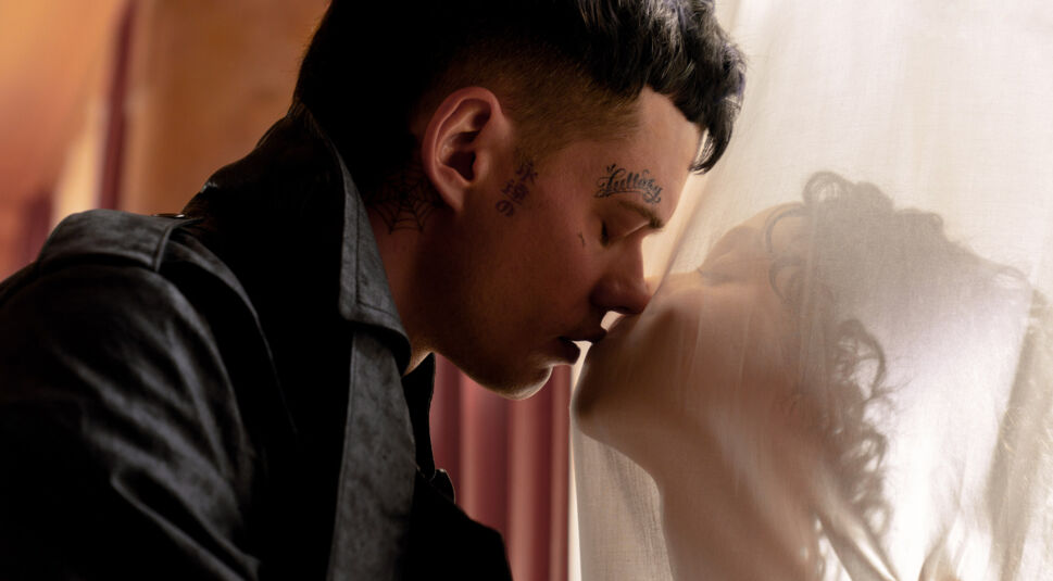 Auf dem Bild zu "The Crow Unterschiede" sind Bill Skarsgård (links) und FKA Twigs (rechts) in einer intimen Szene zu sehen. Skarsgård, mit mehreren Gesichtstattoos, lehnt sich mit geschlossenen Augen nach vorne, während er durch einen halbtransparenten Vorhang FKA Twigs' Gesicht sanft berührt. Ihre Silhouette ist weich durch den Vorhang erkennbar, was der Szene eine geheimnisvolle und zärtliche Atmosphäre verleiht.