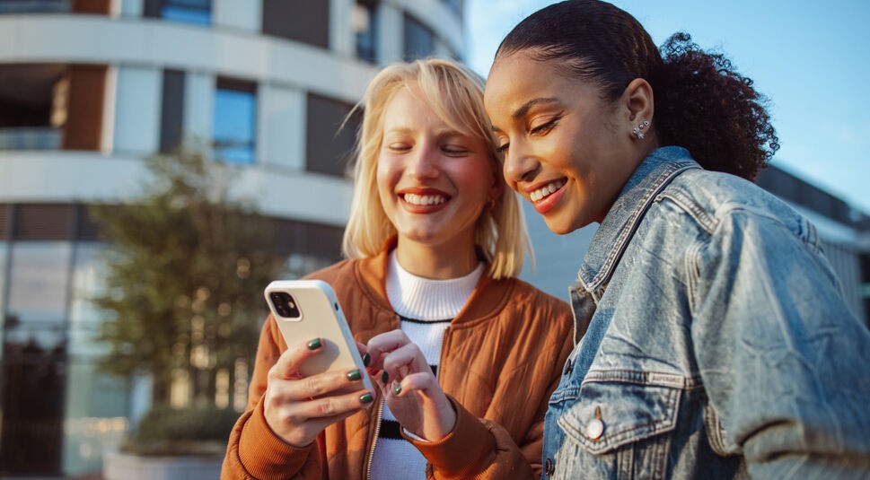 Auf dem Bild in dem Artikel zu Vodafone Young Tarifen lächeln zwei junge Frauen, während sie gemeinsam auf ein Smartphone schauen.
