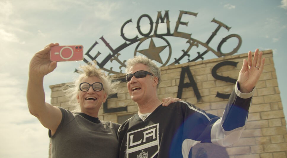 Auf dem Bild zu Will Ferrels-Netflix-Doku-Artikel sind Will Ferrell und Harper Steele zu sehen, wie sie lachend ein Selfie vor einem „Welcome to Texas“-Schild machen. Ferrell trägt ein LA Kings-Trikot und Sonnenbrille, während Steele eine runde Brille und ein graues T-Shirt trägt. Beide wirken ausgelassen und freundlich, als sie in die Kamera lächeln und die Hand heben.