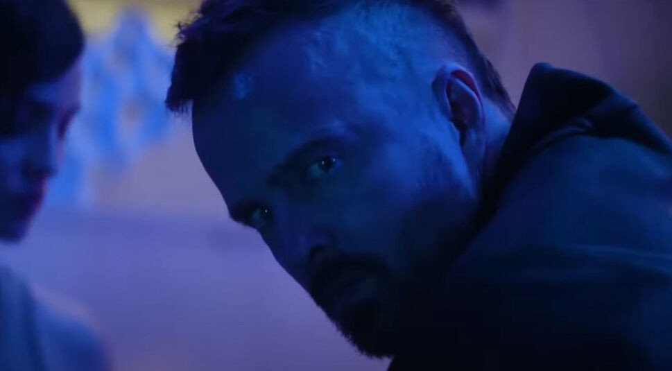 In dieser Szene aus dem Film Ash ist Aaron Paul in einer Nahaufnahme zu sehen. Sein Gesicht ist in ein mysteriöses, bläuliches Licht getaucht, und sein intensiver, ernster Blick ist zur Seite gerichtet, als ob er etwas oder jemanden beobachtet. Im Hintergrund ist eine unscharfe Gestalt zu erkennen, die eine bedrückende und geheimnisvolle Atmosphäre verstärkt.