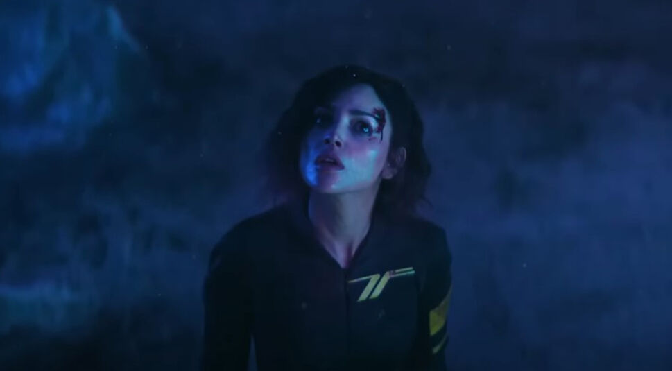 In dieser Nahaufnahme aus dem Film Ash ist Eiza González mit einem verletzten Gesicht zu sehen, ihre Stirn blutet leicht. Ihr Gesichtsausdruck wirkt erschrocken und angespannt, während sie in einem blauen Licht angestrahlt wird, das eine düstere und geheimnisvolle Atmosphäre schafft. Der Hintergrund ist dunkel und undeutlich, was die dramatische Spannung der Szene verstärkt.