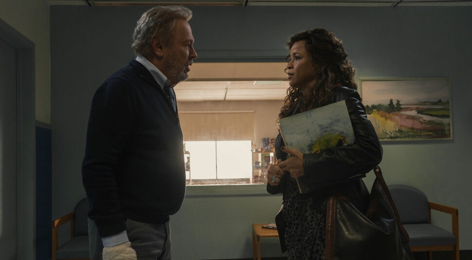 In einer Szene aus der Before-Serie auf Apple TV+ stehen Eli, gespielt von Billy Crystal, und Denise, dargestellt von Rosie Perez, einander gegenüber. Eli trägt ein dunkles Pullover und hält seine bandagierte Hand, während Denise mit einer großen Tasche und einem Ordner in der Hand ernst schaut. Die Spannung zwischen den beiden ist spürbar, da sie sich in einem schlicht eingerichteten Raum mit Kunst an der Wand befinden.