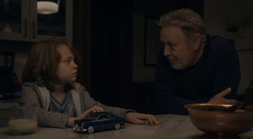 n einer Szene aus der Before-Serie auf Apple TV+ sind Eli, gespielt von Billy Crystal, und Noah, dargestellt von Jacobi Jupe, an einem Küchentisch zu sehen. Noah sitzt links mit einem Modellauto vor sich und einem Glas Milch daneben, während Eli rechts ihm ruhig in die Augen schaut. Die Atmosphäre ist nachdenklich und ruhig, und die Szene deutet auf eine tiefere Verbindung zwischen den beiden Charakteren hin.