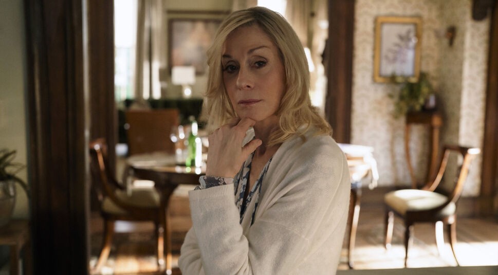 In dieser Szene aus der Before-Serie auf Apple TV+ ist Lynn, gespielt von Judith Light, in einem elegant eingerichteten Raum zu sehen. Sie trägt einen hellen Cardigan und schaut nachdenklich in die Kamera, während sie eine Hand an ihr Kinn legt. Im Hintergrund ist ein stilvoller Essbereich mit Holzstühlen und einer Flasche auf dem Tisch zu erkennen, was eine ruhige und reflektierte Stimmung vermittelt.