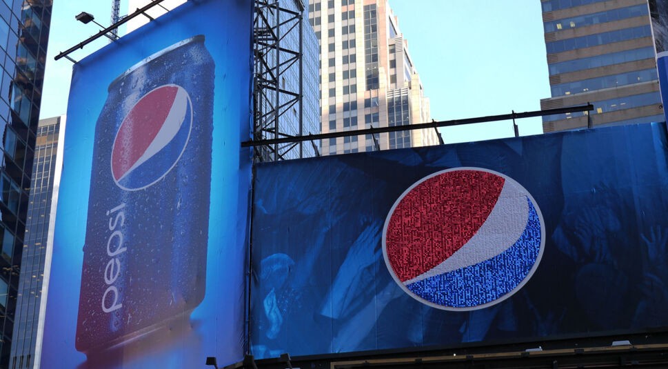 Auf diesem Bild zur "Cola Wars: Wahre Geschichte" sind zwei große Pepsi-Plakatwerbungen in einer urbanen Umgebung zu sehen. Links ist eine eisgekühlte Pepsi-Dose mit Wassertropfen abgebildet, während rechts ein überdimensionales Pepsi-Logo auf einem weiteren Plakat zu sehen ist. Die Szenerie zeigt moderne Hochhäuser im Hintergrund, was die starke Präsenz von Pepsi in der Stadtlandschaft unterstreicht und symbolisch für den Werbekampf in den Cola Wars steht.