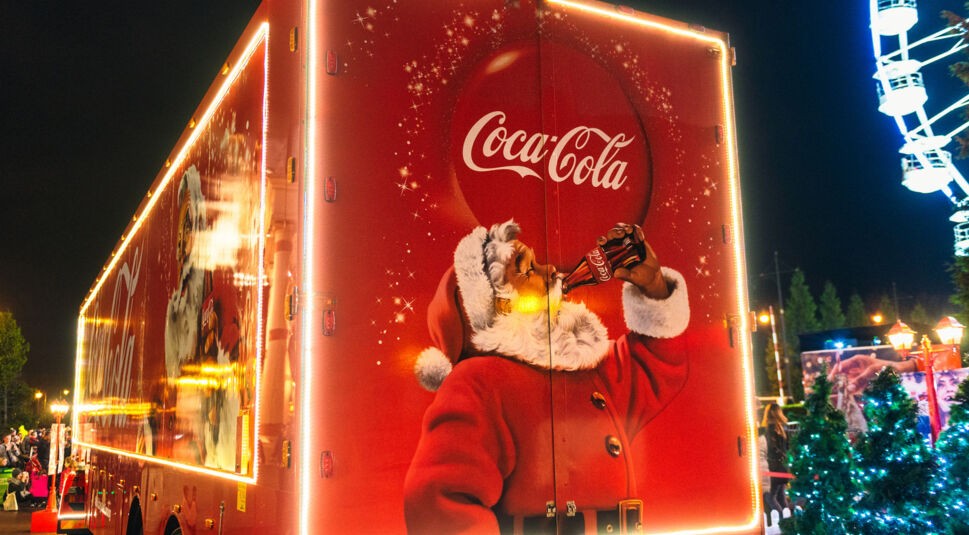 Auf diesem Bild zur "Cola Wars: Wahre Geschichte" ist ein festlich beleuchteter Coca-Cola-Truck zu sehen, auf dem der ikonische Weihnachtsmann eine Flasche Coca-Cola genießt. Der Truck ist in leuchtendem Rot gehalten und mit weißen Lichtern umrahmt, was eine weihnachtliche Stimmung erzeugt. Im Hintergrund ist ein beleuchtetes Riesenrad und geschmückte Tannenbäume zu erkennen, die die festliche Atmosphäre verstärken. Der Weihnachtsmann auf dem Truck symbolisiert die berühmte Coca-Cola-Weihnachtskampagne, die Teil des Marketingwettbewerbs im Cola-Krieg ist.