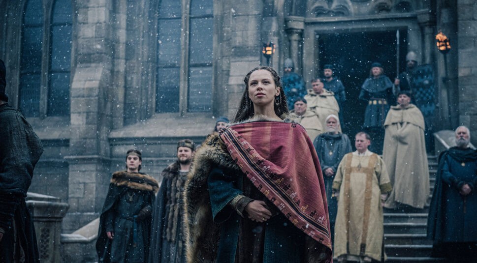 In dieser Szene aus "Hagen Im Tal der Nibelungen" steht Kriemhild, die Königstochter, im Mittelpunkt vor einer großen, steinernen Kirche. Sie trägt einen prachtvollen Umhang mit rotem Stoff und Pelzbesatz und blickt stolz und entschlossen in die Ferne. Hinter ihr stehen Adlige und Geistliche auf den Stufen der Kirche, einige mit ernsten Gesichtern. Schneeflocken fallen sanft vom Himmel und verstärken die feierliche und dramatische Stimmung der Szene. Die historische Kleidung und die imposante Architektur unterstreichen die mittelalterliche Atmosphäre.