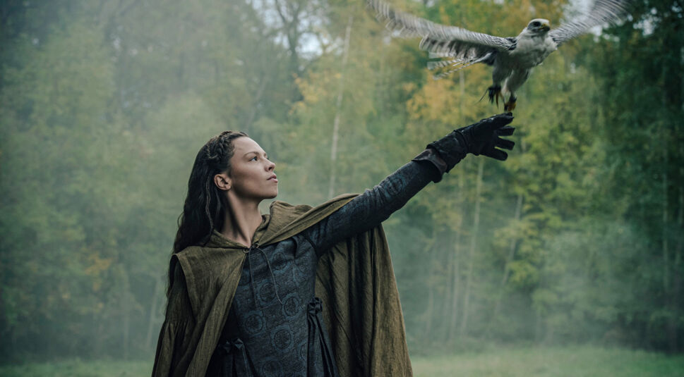 Auf dem Bild aus "Hagen – Im Tal der Nibelungen" ist Kriemhild, gespielt von Lilja van der Zwaag, zu sehen. Sie trägt ein dunkles, gemustertes Gewand und einen braunen Umhang, während sie einen Falken auf ihrer behandschuhten Hand fliegen lässt. Die Szene spielt in einem nebligen Wald, der die mystische und naturverbundene Atmosphäre verstärkt.