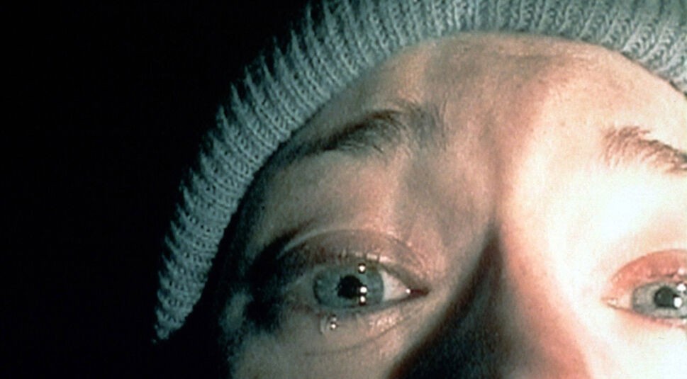 Das Bild im Artikel zu Horrorfilm-Anekdoten zeigt eine ikonische Nahaufnahme aus dem Film Blair Witch Project, in der eine erschrockene Person mit weit aufgerissenen, tränengefüllten Augen und einem Wollhut zu sehen ist, eingefangen in einer düsteren Atmosphäre.