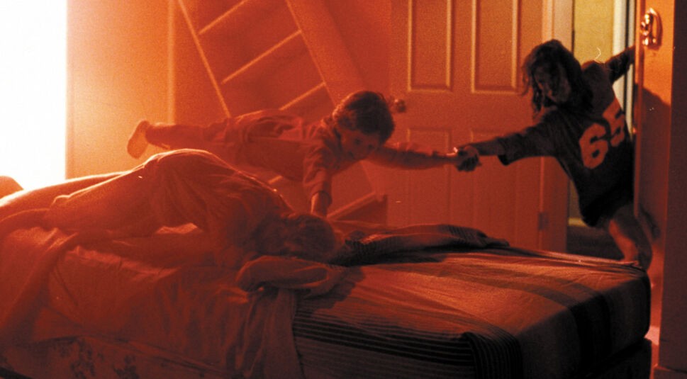 Das Bild im Artikel zu Horrorfilm-Anekdoten zeigt eine Szene aus dem Film Poltergeist, in der zwei Kinder in einem orange-rötlich beleuchteten Raum scheinbar von einer unsichtbaren Kraft angezogen werden. Eines der Kinder hält sich an der Tür fest, während das andere in der Luft schwebt und versucht, ein drittes Kind auf dem Bett zu erreichen. 