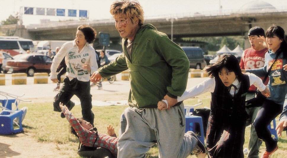 Das Bild im Artikel zu Horrorfilm-Anekdoten zeigt eine Szene aus dem Film The Host, in der mehrere Menschen panisch weglaufen. Im Vordergrund hält ein Mann in grüner Jacke die Hand eines Mädchens, während sie vor einer Bedrohung fliehen. 