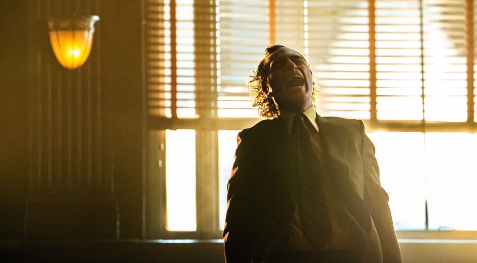 Auf dem Bild zu „Joker: Folie à Deux Ende erklärt“ sieht man den Joker, gespielt von Joaquin Phoenix, in einem dramatischen Moment, wie er lauthals lacht. Er steht in einem Raum mit dunklem Holzinterieur, beleuchtet durch das warme Licht der tief stehenden Sonne, die durch Jalousien fällt. Der Joker trägt einen dunklen Anzug und seine Haare sind zerzaust. Sein Lachen und die starke Gegenlichtstimmung verleihen der Szene eine intensive und unheimliche Atmosphäre.