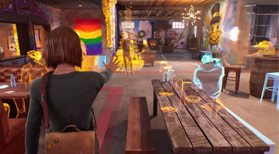 Auf dem Bild aus Life is Strange: Double Exposure Test ist Max inmitten einer Szene zu sehen, in der sie ihre besondere Fähigkeit, den „Puls“, einsetzt. Um sie herum erscheinen schemenhafte, farbige Umrisse von Personen und Gegenständen in Orange und Blau, als ob sie in eine andere Realität oder Erinnerung eintaucht. Max steht mit ausgestrecktem Arm in einem Raum, dekoriert mit einer Regenbogenflagge und Graffiti an den Wänden. Die Atmosphäre wirkt mystisch und surreal.