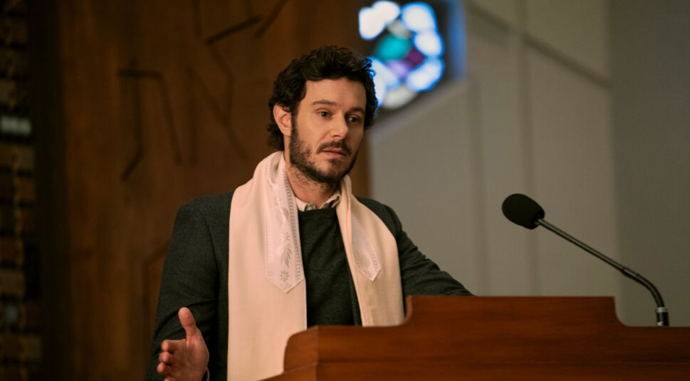 Auf dem Bild zur Serie „Nobody Wants This“ ist Noah, gespielt von Adam Brody, zu sehen, wie er an einem Rednerpult steht und spricht. Er trägt einen dunklen Anzug und einen hellen Schal, der ihn in einem formellen, möglicherweise religiösen Kontext zeigt. Noah wirkt nachdenklich, während er gestikuliert und seine Rede hält. Im Hintergrund sind die unscharfen Farben eines Kirchenfensters zu erkennen, was die ernste und reflektierende Atmosphäre der Szene unterstreicht.