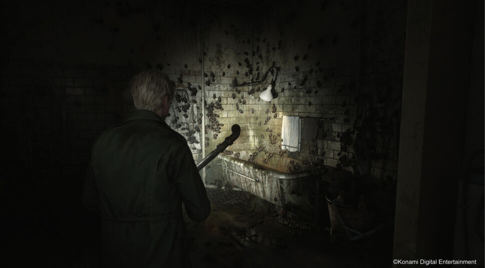 In diesem Bild aus dem "Silent Hill 2 Remake im Test" steht James Sunderland in einem verfallenen Badezimmer. Er trägt einen grünen Mantel und hält eine Eisenstange in der Hand. Die Wände und die schmutzige Badewanne sind von Kakerlaken und Dreck bedeckt, wodurch die düstere und bedrückende Atmosphäre verstärkt wird. Der Raum ist schwach beleuchtet, was die unheimliche Stimmung und das Gefühl der Bedrohung im Spiel unterstreicht.
