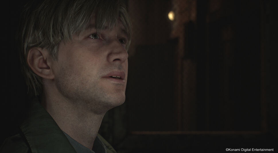 In diesem Bild aus dem "Silent Hill 2 Remake im Test" ist eine Nahaufnahme von James Sunderland zu sehen. Er blickt nachdenklich nach oben, sein Gesichtsausdruck wirkt ernst und leicht melancholisch. Die schwache Beleuchtung im Hintergrund, die dunklen Schatten und das subtile Licht auf seinem Gesicht betonen die emotionale und düstere Atmosphäre des Spiels. Sein blonder, leicht zerzauster Haarschnitt und der olivgrüne Mantel sind deutlich erkennbar.